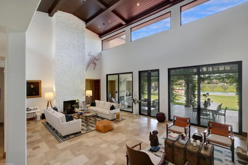 Double-height great room with a stone chimney, exposed timber ceiling and glazing onto the lawn